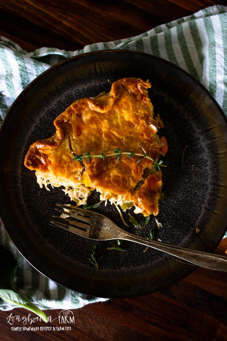 A slice of golden, flaky homemade chicken pot pie garnished with herbs sits on a dark plate with a fork, atop a striped green and white cloth on a wooden table.