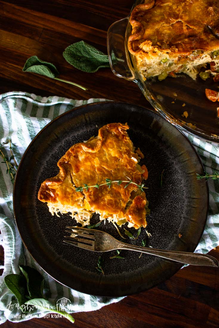 A slice of golden-brown homemade chicken pot pie garnished with herbs sits on a dark plate with a fork, next to a striped cloth. The remaining pie is in a glass dish nearby on a wooden table.