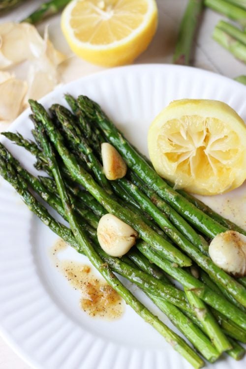 Roasted asparagus on a plate topped with roasted garlic cloves next to a lemon half.