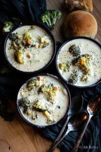 three black bowls of broccoli cheddar soup with bread rolls to the side