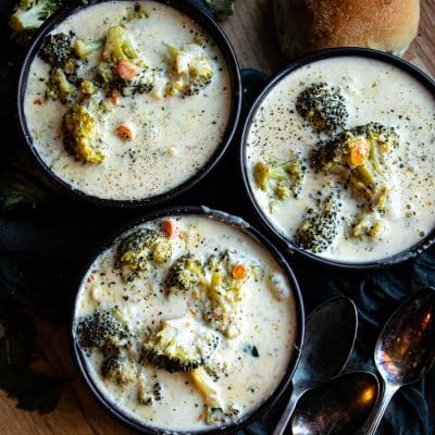 three black bowls of broccoli cheddar soup with bread rolls to the side