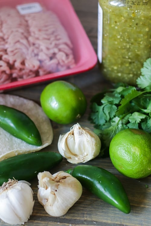 Garlic cloves, limes, jalapeños, and cilantro next to a package of ground pork and jar of salsa verde.