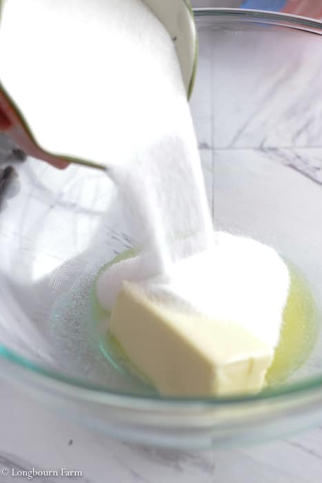 Sugar being poured over a stick of butter in a glass bowl.