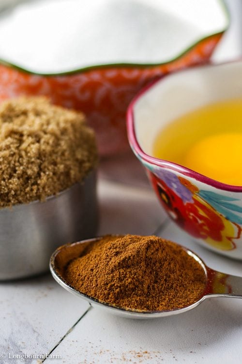 Measuring cup with white sugar next to a measuring cup with brown sugar, next to a bowl with cracked eggs next to a heaping tablespoon of cinnamon on a countertop.