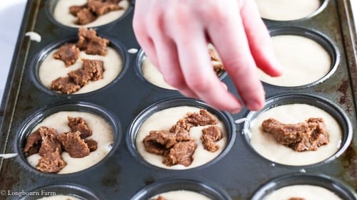 A hand placing clumps of the strussel topping onto the cinnamon muffin batter already portioned in a muffin tin.