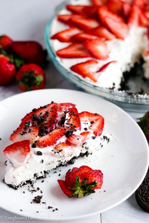 Slice of no bake strawberry cheesecake on a plate with strawberries and Oreos nearby with the whole cheesecake in the background. 