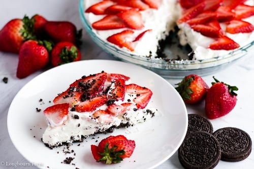 Horizontal image of a slice of no bake strawberry cheesecake on a plate with strawberries and Oreos nearby with the whole cheesecake in the background. 