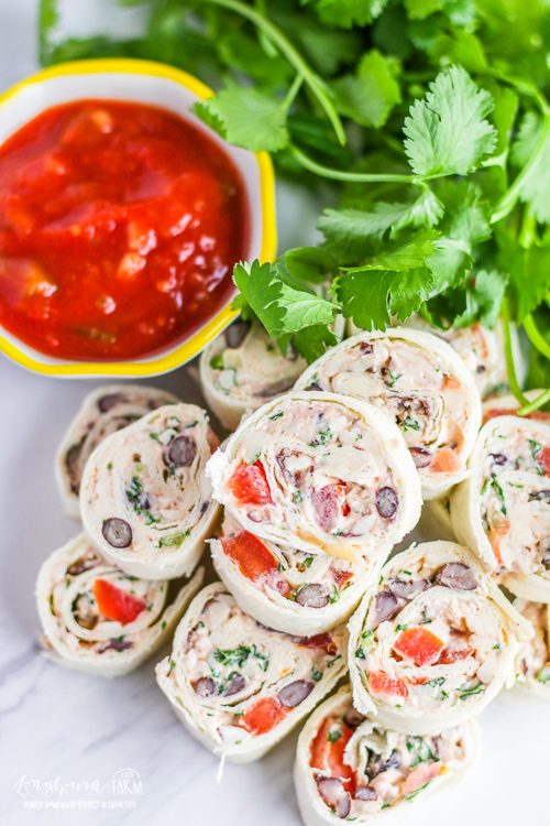 Top view of stack of tex-mex tortilla pinwheels with a cup of salsa next to it and cilantro in the background. 