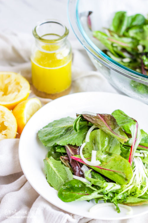 Easy green salad on a white plate next to the salad bowl, lemons and lemon vinaigrette.