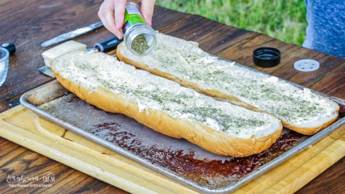 Sprinkling Italian seasoning on buttered french bread for cheesy garlic bread.