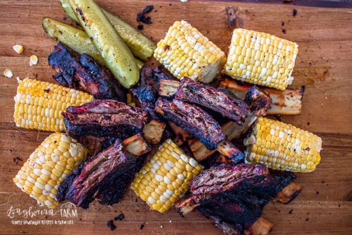 Smoked beef ribs piled on a cutting board with corn on the cob. 