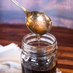 a glass jar with syrup inside below a spoon thats drizzling more syrup into the jar