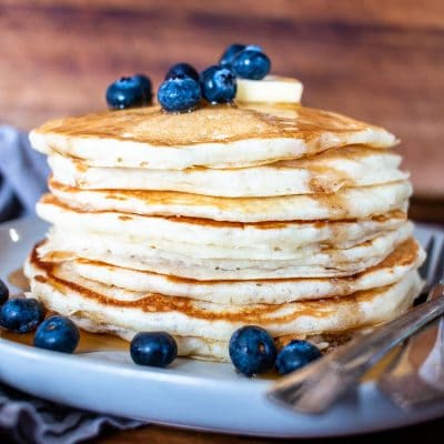 a upclose side view of a stack of pancakes on a plate with syrup, fresh blueberries, a slab of butter and two forks
