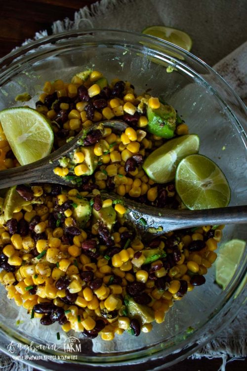 lime wedges and wooden spoons in a glass bowl full of black beans, corn, and avocado sald