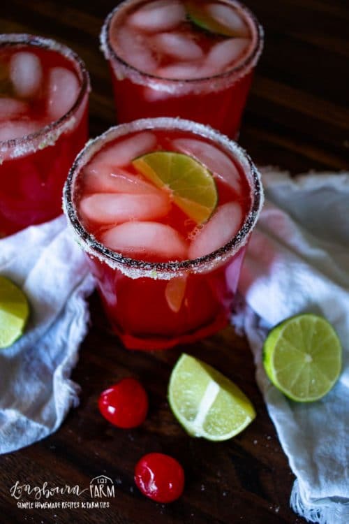 lime and cheeries around cups of sugar rimmed cherry limeade