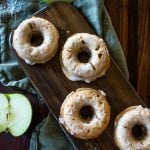 glazed apple cider donuts on a long wooden board with fresh apple slices to the sides