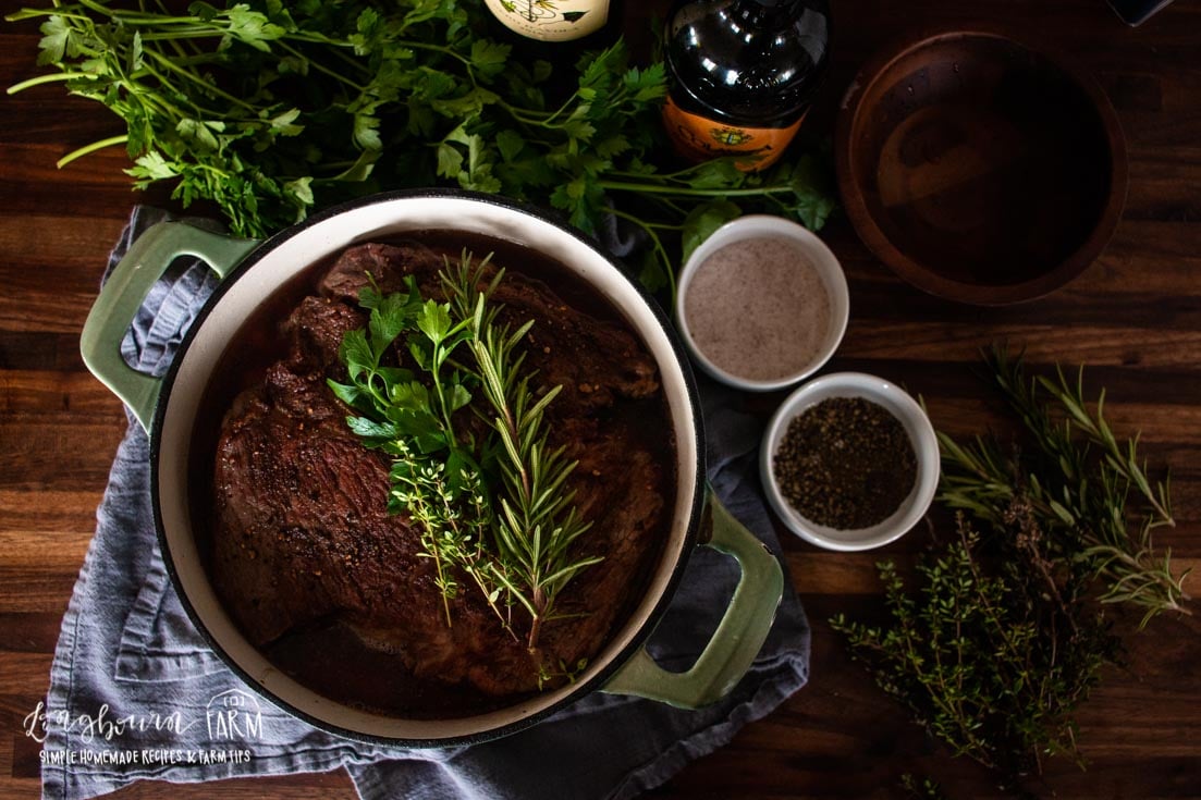 a dutch oven filled with cooked chuck roast and more ingredients off to the sides with fresh herbs on top
