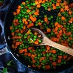 an upclose aerial view of a wooden spoon in a skillet of cooked peas and carrots