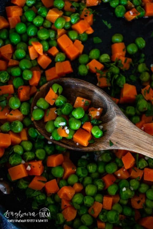 an upclose view of a wooden spoon filled with cooked peas and carrots