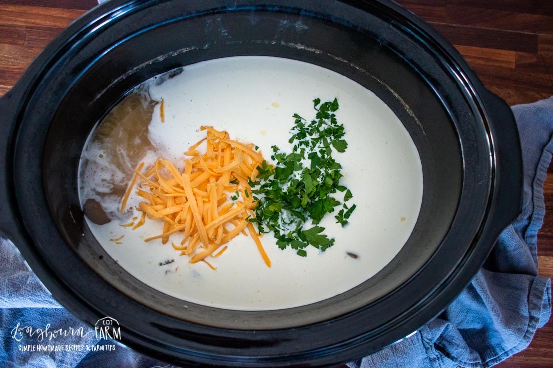 shredded cheese and chopped herbs being added to the crockpot full of ham soup and milk