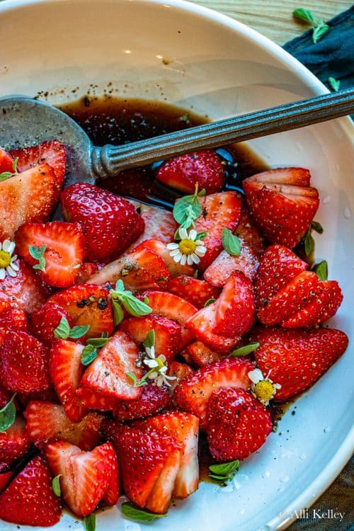 a bowl of balsamic strawberries garnished with fresh herbs and daisies with a spoon