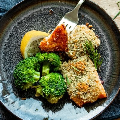 a fork taking some salmon with a herbed crust, broccoli and lemon wedge on a plate
