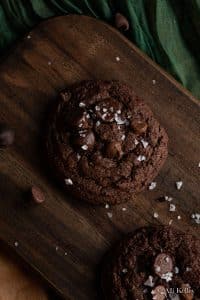 double chocolate chip cookies on a wooden chopping board with sea salt flakes sprinkled on top.