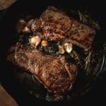 two new york steaks cooking in a pan on the stovetop with fresh herbs, garlic and butter