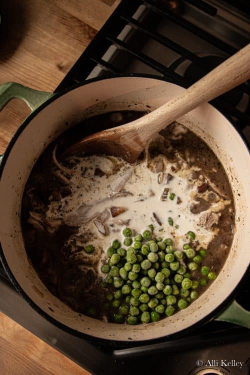 a wooden spoon about to mix chicken stew, milk and frozen peas together in a large pan on the stovetop