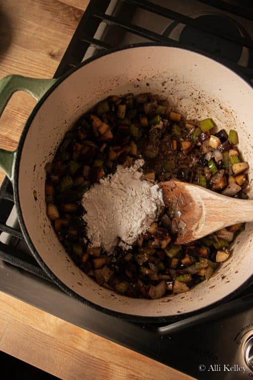 a wooden spoon mixing flour with cooked purple carrots, onions and celery in a large pan on the stovetop