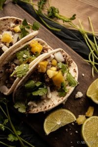 ground pork tacos with chopped pineapple, onions, fresh cilantro and cheese on a wooden chopping board with lime wedges
