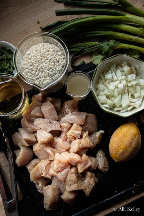 chunks of raw chicken, a lemon, a bowl of chopped onions, a jug of chicken broth and white wine, a jug of risotto rice, fresh herbs and whole green onions on a slate clopping board