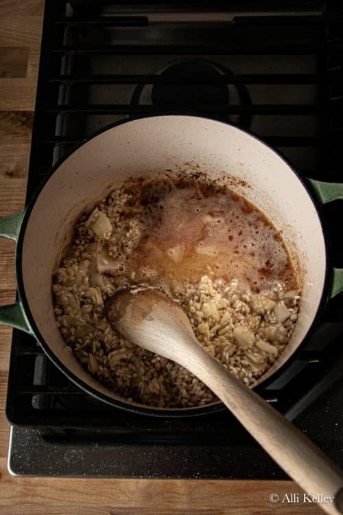 a wooden spoon mixing uncooked risotto rice and sliced shallot in a pan