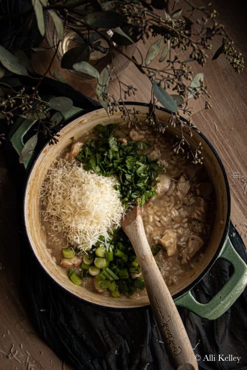 a wooden spoon about to mix chicken risotto with fresh herbs, chopped green onions and parmesan cheese in a pan