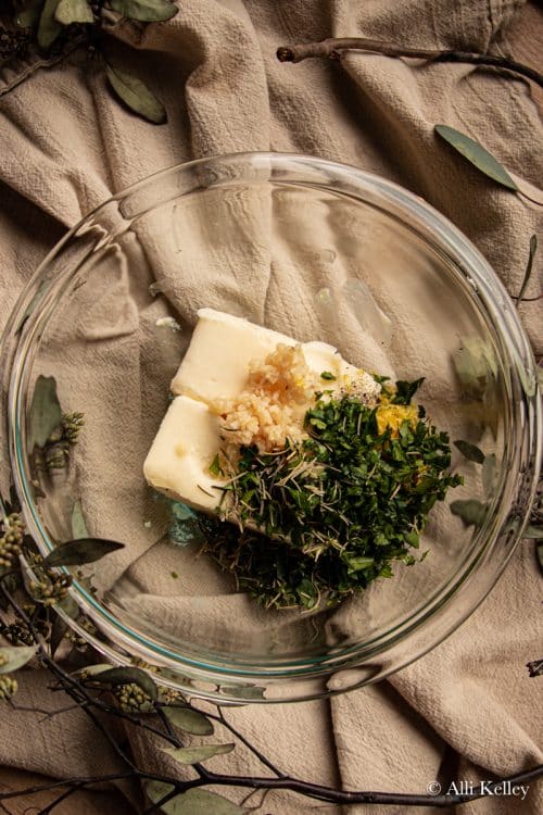 two sticks of butter, fresh herbs and garlic in a glass mixing bowl