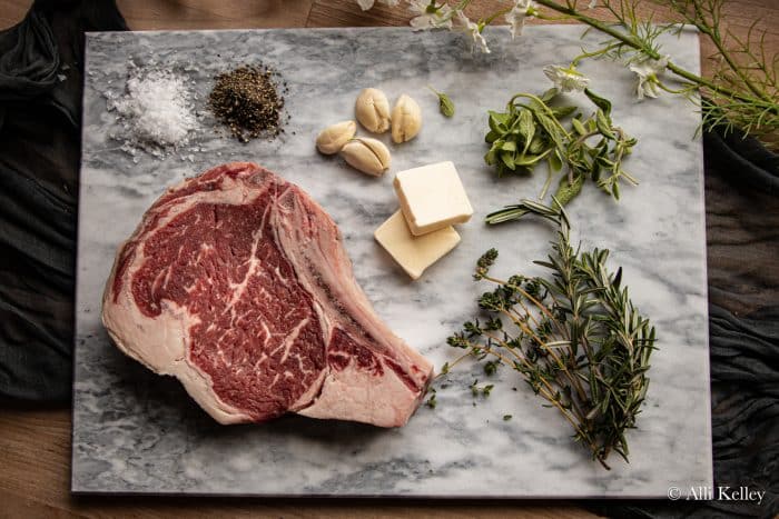 raw cowboy steak with seasonings, fresh herbs, garlic cloves, and butter on a chopping board
