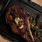 cowboy steak on a chopping board with herbs and garlic cloves
