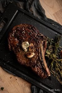 cowboy steak on a chopping board with herbs and garlic cloves