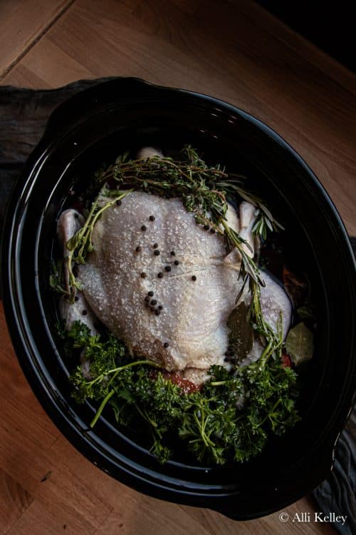 a whole uncooked chicken topped with seasonings and fresh herbs in a crock pot