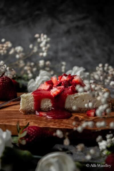 a slice of vanilla cheesecake, topped with strawberry glaze and fresh strawberry slices on a wooden chopping board