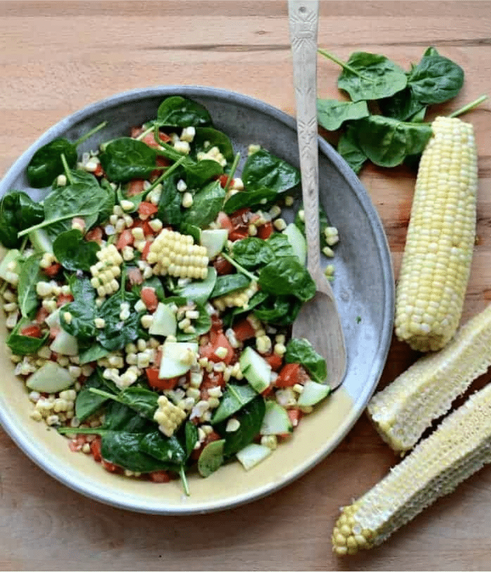 A vibrant corn spinach salad with tomatoes and cucumber sits in a bowl on a wooden surface beside three ears of corn, one partially husked. A wooden spoon rests in the bowl, ready to serve.