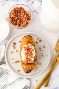 An air fryer hasselback potato on a plate topped with sour cream, cheese sauce, and bacon bits sits next to a bowl of extra bacon bits, a fork and knife, and a striped napkin on a marble surface.