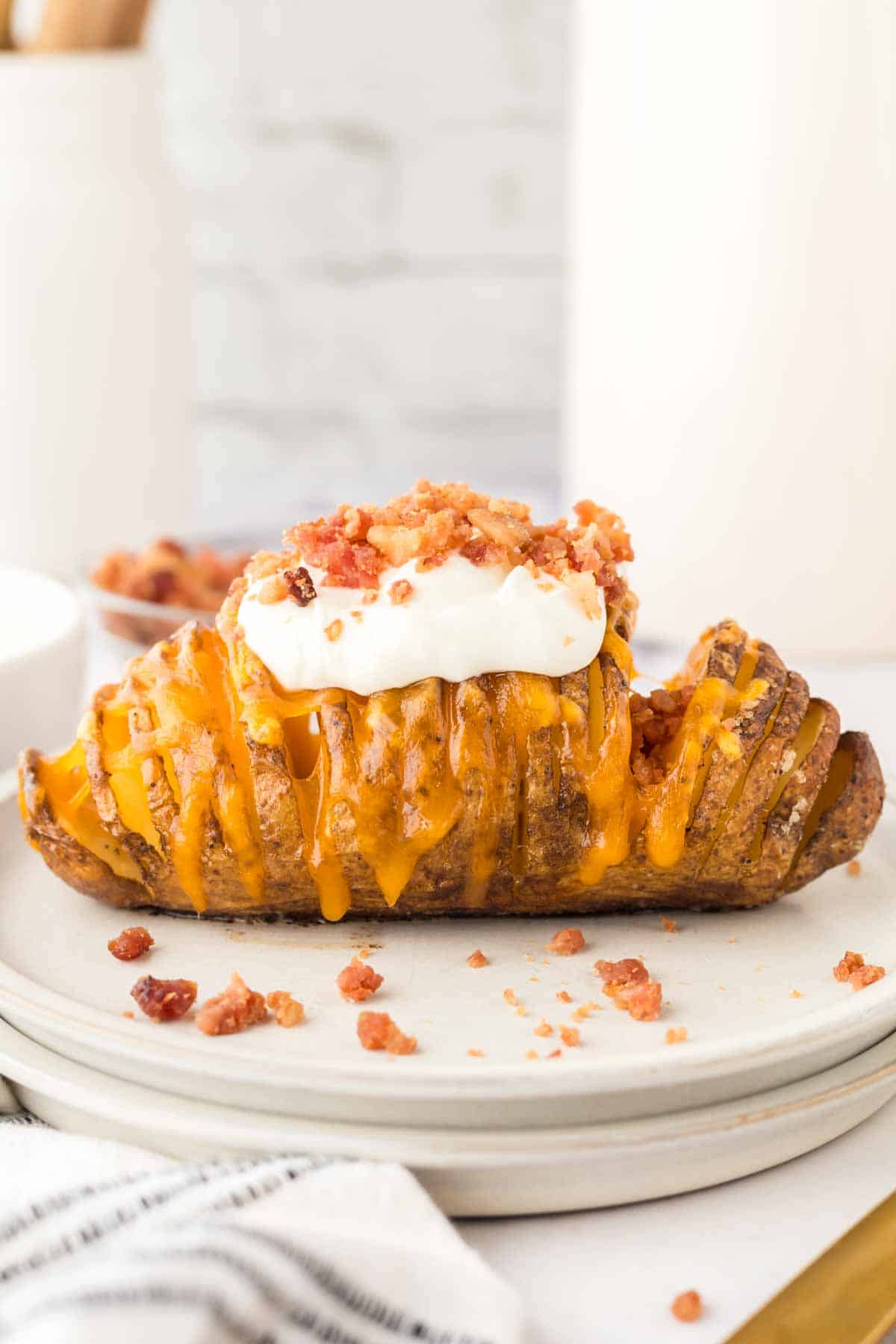 An air fryer Hasselback baked potato with melted cheddar cheese, topped with a dollop of sour cream and crispy bacon bits, served on a white plate.