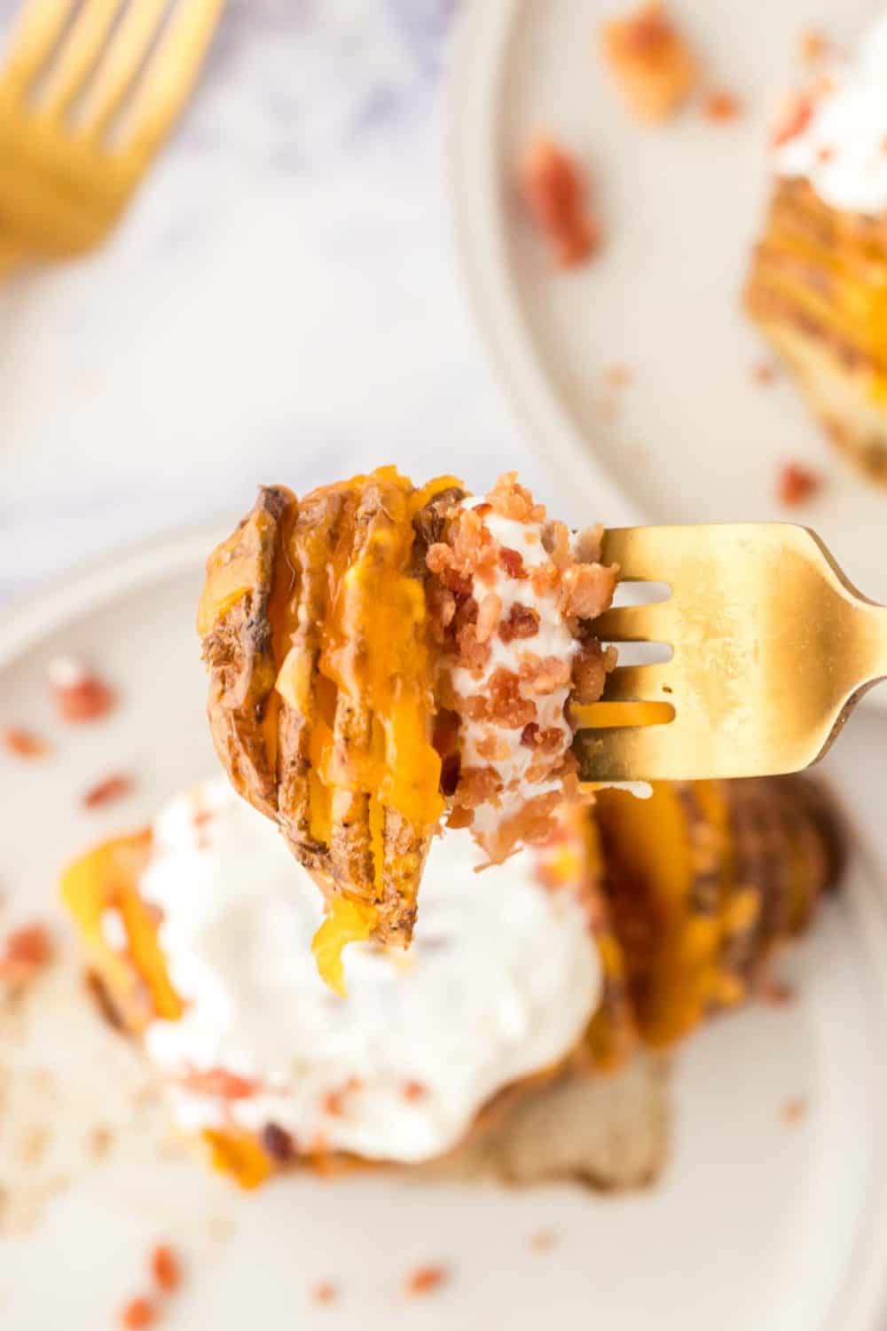 A close-up of a fork holding a bite of loaded sweet potato topped with melted cheese, sour cream, and bacon bits, with air fryer hasselback potatoes served on a plate in the background.