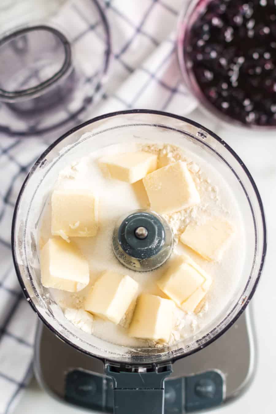 Overhead view of a food processor containing cubes of butter, flour, and sugar—ingredients for blueberry pie bars—with a bowl of dark berries and a checkered cloth visible in the background.