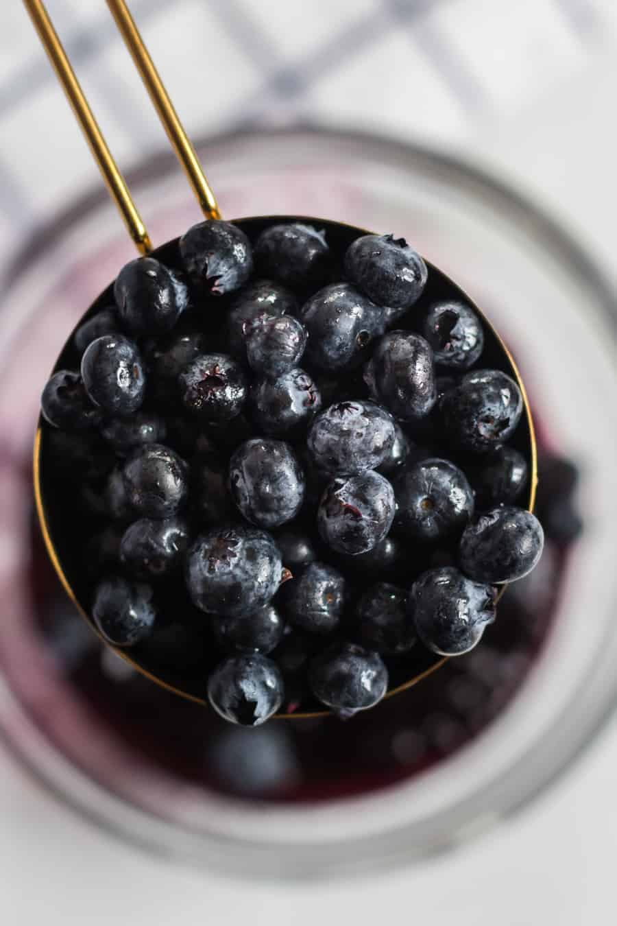 A close-up of fresh blueberries piled in a gold measuring cup, ready to be used for delicious blueberry pie bars, held above a glass bowl with a blurred background.