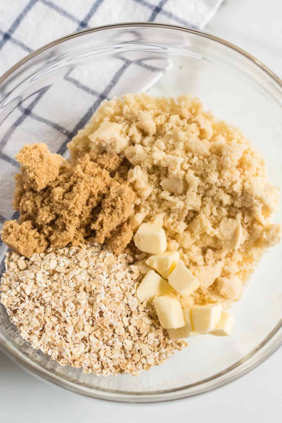 A glass bowl containing brown sugar, crumbly flour mixture, chopped butter, and rolled oats—ingredients perfect for blueberry pie bars—sits on a white surface with a checkered cloth in the background.