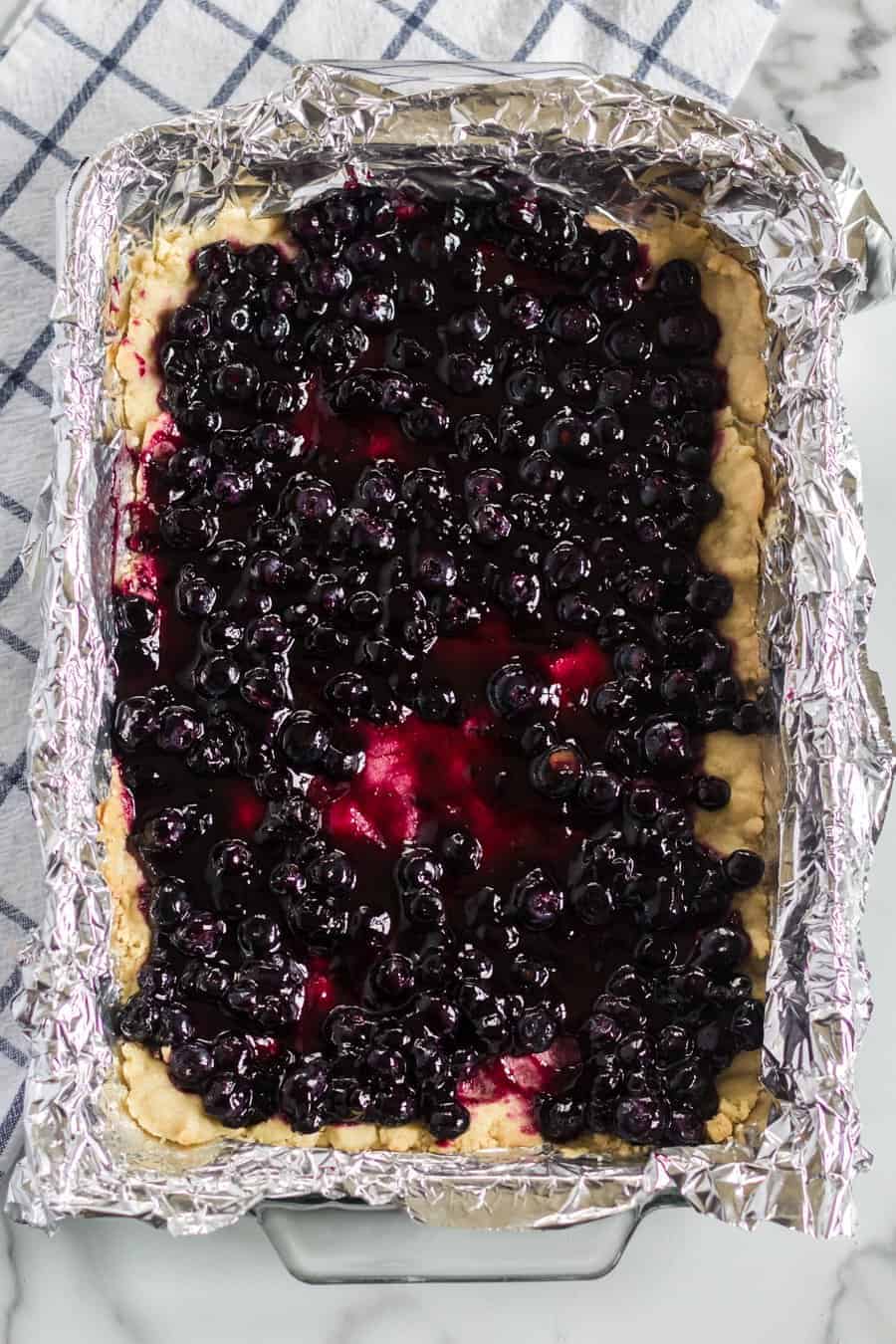 A rectangular baking dish lined with foil holds a partially filled dessert with a golden crust and a layer of glossy blueberry topping. A checked kitchen towel is in the background on a marble surface.