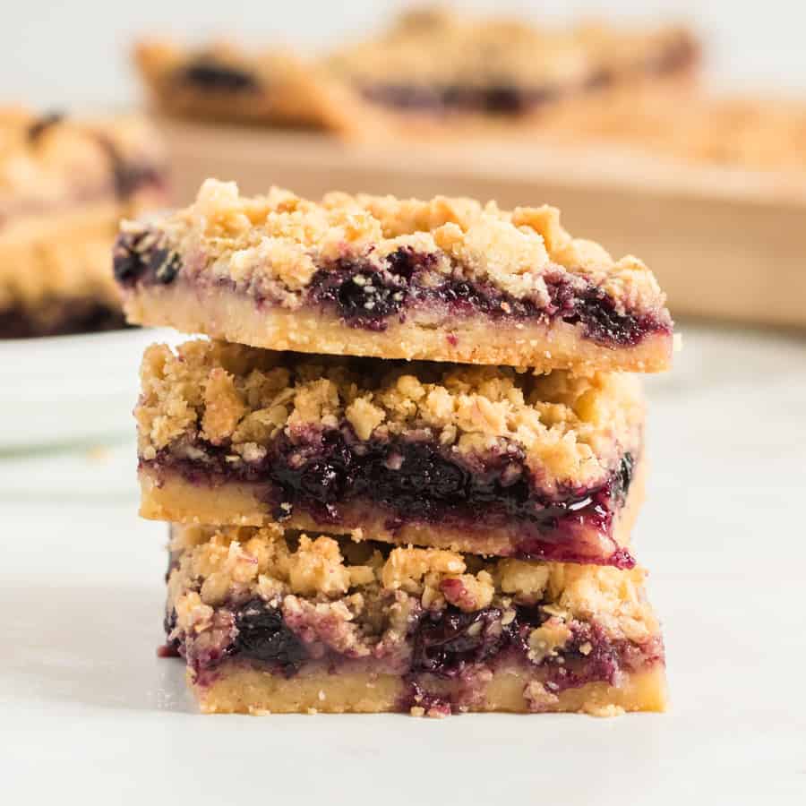 A stack of three crumbly blueberry pie bars with a golden oat topping reveals layers of blueberry filling and buttery crust, sitting on a white surface with more bars blurred in the background.