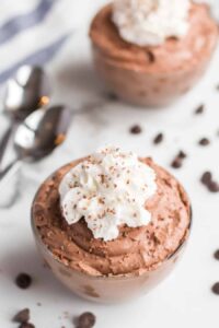 A glass bowl filled with creamy chocolate mousse, topped with a swirl of whipped cream and sprinkled chocolate shavings. In the background, hints of another bowl tease a tempting chocolate mousse recipe waiting to be enjoyed.